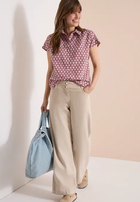 Frau trägt rosa gemustertes Oberteil, beige weite Hose und hält großen hellblauen Shopper.